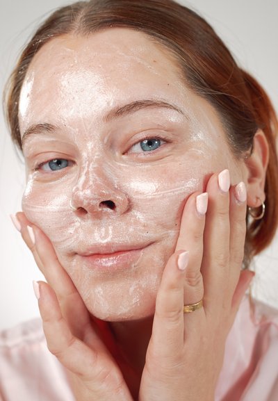 Primer plano de un rostro con una mascarilla facial ligera y espumosa. Piel suave y luminosa y uñas de color rosa suave, con manos que tocan suavemente el rostro.