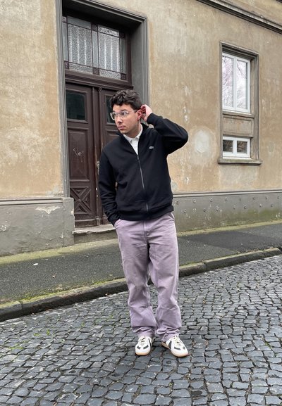 Chaqueta negra con cremallera, camisa blanca con cuello, pantalones lilas claros. Gafas transparentes, zapatillas neutras, de pie en una calle de adoquines cerca de una puerta.