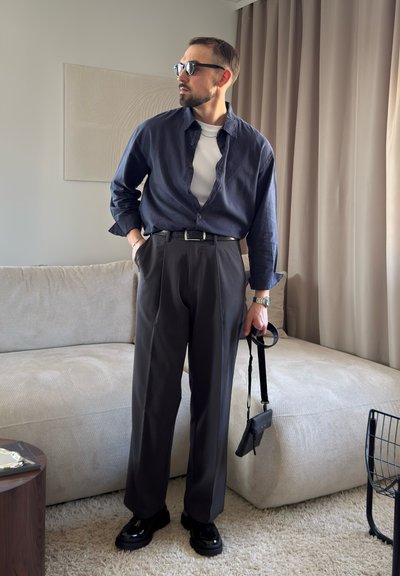 Hombre con gafas de sol, camisa azul oscuro sobre camiseta blanca, pantalones grises, zapatos negros, sosteniendo una pequeña bolsa negra, de pie en una sala de estar beige.