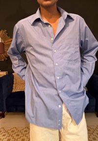 Άνδρας που φοράει ανοιχτό γαλάζιο πουκάμισο με κουμπιά και μπεζ παντελόνι, στέκεται σε εσωτερικό χώρο με τα χέρια πίσω από την πλάτη.