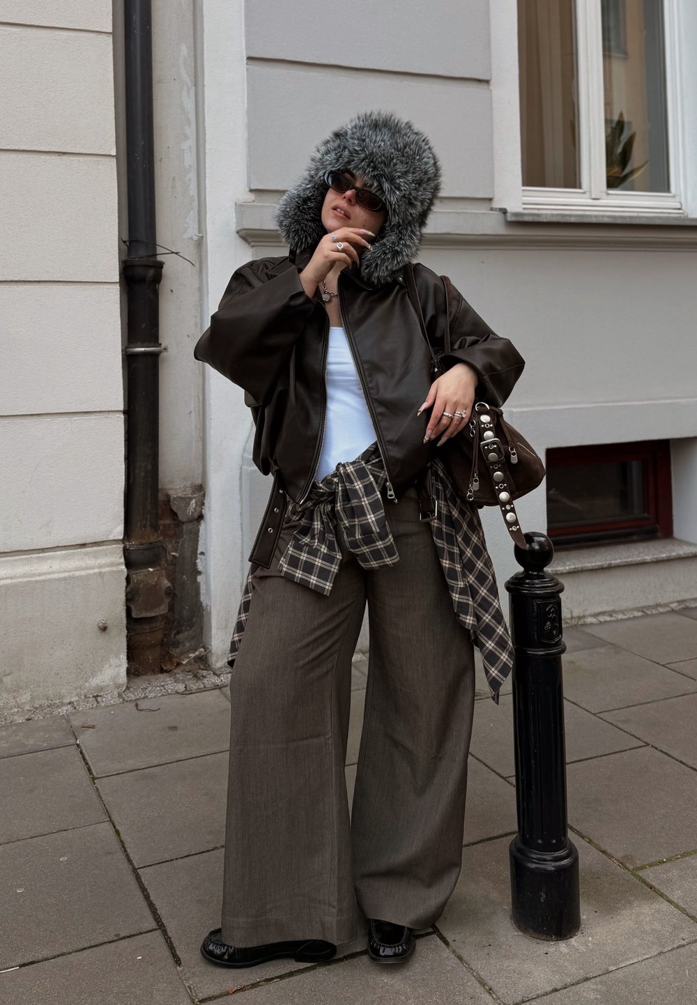 Giacchetto in pelle marrone con cappuccio in pelliccia sintetica, canottiera bianca, camicia a quadri annodata in vita, pantaloni beige larghi e scarpe nere.