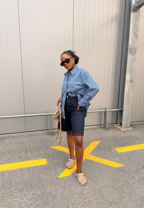 Frau mit Sonnenbrille, blau kariertem Hemd, dunklen Shorts und beigen Clogs, die auf gelben Fahrbahnmarkierungen an einer grauen Wand steht und eine beige Handtasche hält.