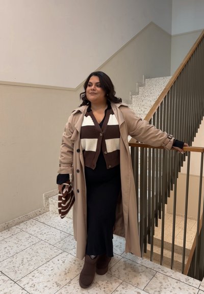 Trench beige sobre un cárdigan a rayas marrón y blanco, combinado con un vestido negro. Botines marrones y un bolso a rayas completan el look.