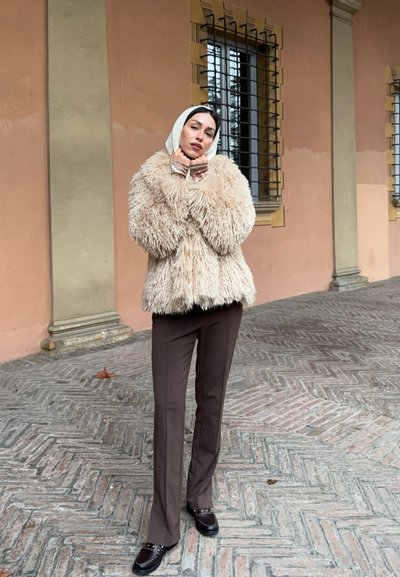 Mujer de pie sobre un pavimento de piedra con diseño, vistiendo un abrigo beige de pelo, pantalones marrones, zapatos negros con tachuelas y un pañuelo blanco en la cabeza.
