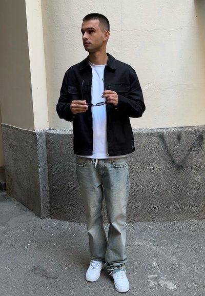 Chaqueta negra, camiseta blanca, jeans de mezclilla azul claro y zapatillas blancas. Sosteniendo gafas de sol, de pie contra una pared texturizada.