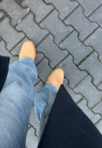 Κίτρινες σουέτ μπότες, μπλε τζην παντελόνι και μαύρο μπουφάν, όπως φαίνονται από ψηλά σε μια ολισθηρή γκρι τοιχοποιία.