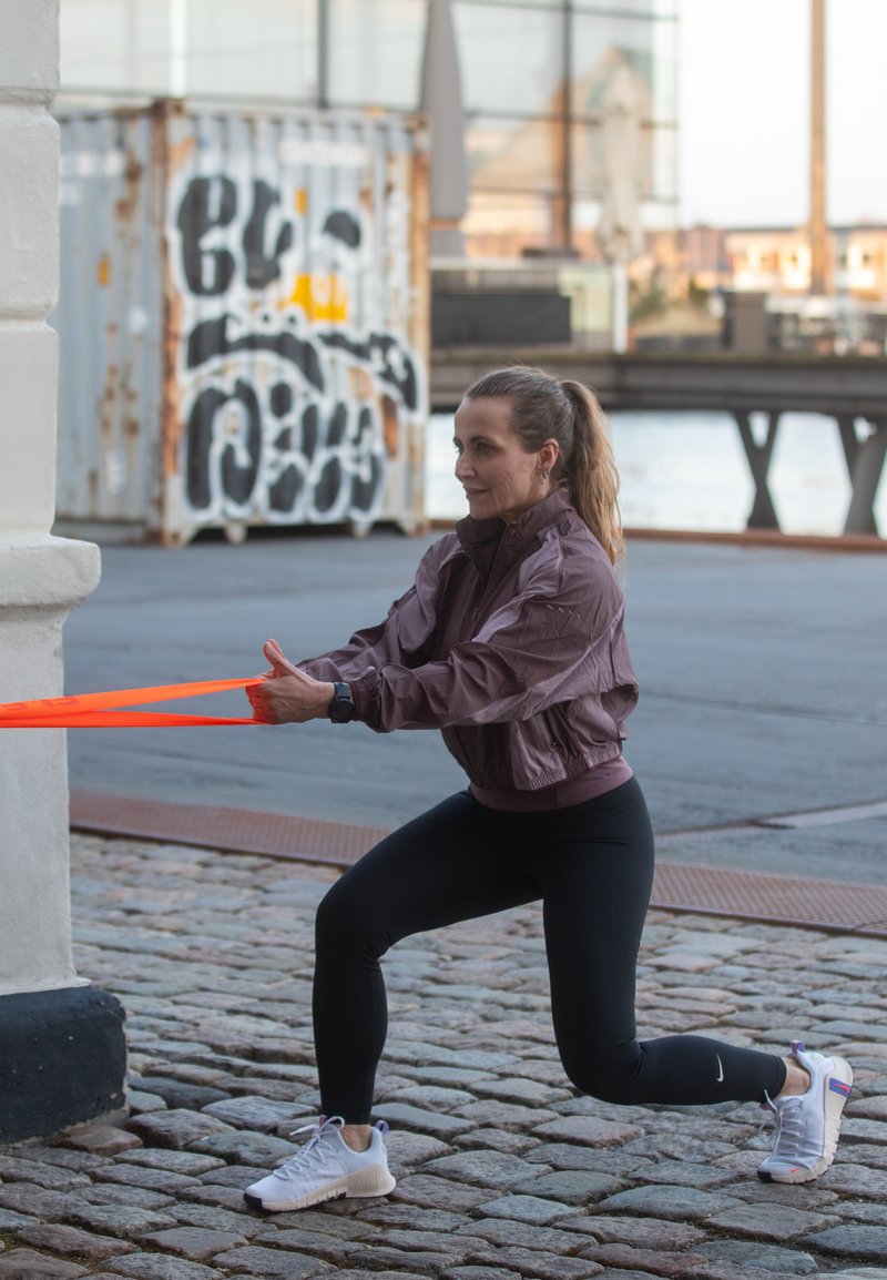 Femme en tenue de sport fait des fentes dans une rue pavée en tirant une bande de résistance orange fixée à un mur près d’un conteneur couvert de graffitis.