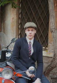 Jauns vīrietis melnā adītā džemperī un raibajā kaklasaitē sēž uz sarkanā skūtera, valkājot pelēku plakanās cepures cepuri pret tekstūrētu akmens sienu.