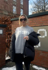 Femme portant des lunettes de soleil foncées, pull gris avec le texte "HORSTIBHV", tenant une doudoune noire, debout près d'un mur en briques en plein air avec des plaques de neige.