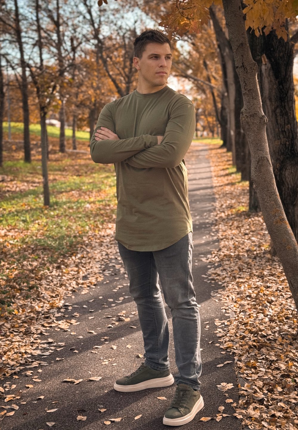 Ελαιόχρουν πουκάμισο με μακριά μανίκια, γκρι εφαρμοστά τζιν και σκούρα πράσινα αθλητικά παπούτσια. Το φόντο περιλαμβάνει ένα μονοπάτι με φυλλωσιά φθινοπώρου και δέντρα.