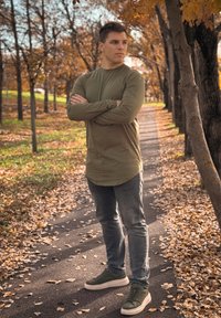 Ελαιόχρουν πουκάμισο με μακριά μανίκια, γκρι εφαρμοστά τζιν και σκούρα πράσινα αθλητικά παπούτσια. Το φόντο περιλαμβάνει ένα μονοπάτι με φυλλωσιά φθινοπώρου και δέντρα.