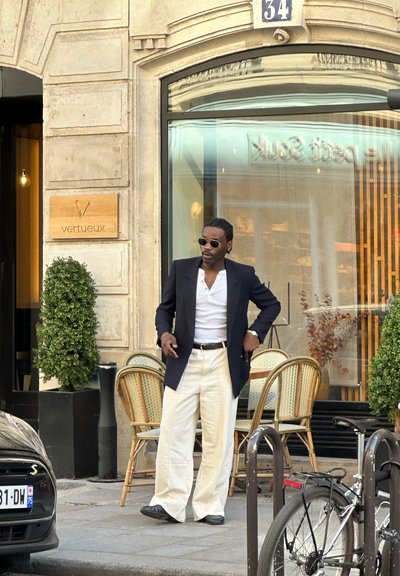 Hombre con gafas de sol, camisa blanca, blazer negro y pantalones blancos, de pie junto a sillas de cafetería fuera del edificio número 34, con una bicicleta y un coche cerca.