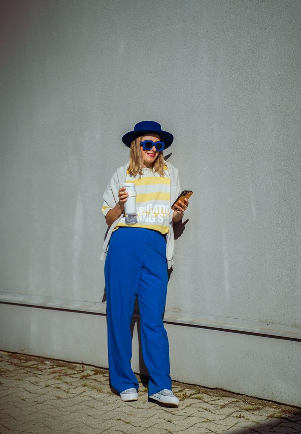 Bluo weite Hosen, übergroßes T-Shirt mit gelb-weißen Streifen, graue Oberbekleidung, blaue Sonnenbrille, blauer Hut, hält ein Getränk und ein Telefon.