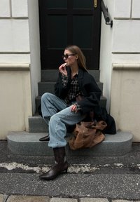 Femme portant des lunettes de soleil, une veste noire, une chemise à carreaux et des bottes, assise sur des marches en pierre près d'une porte sombre, mangeant une collation, avec un sac marron à côté d'elle.