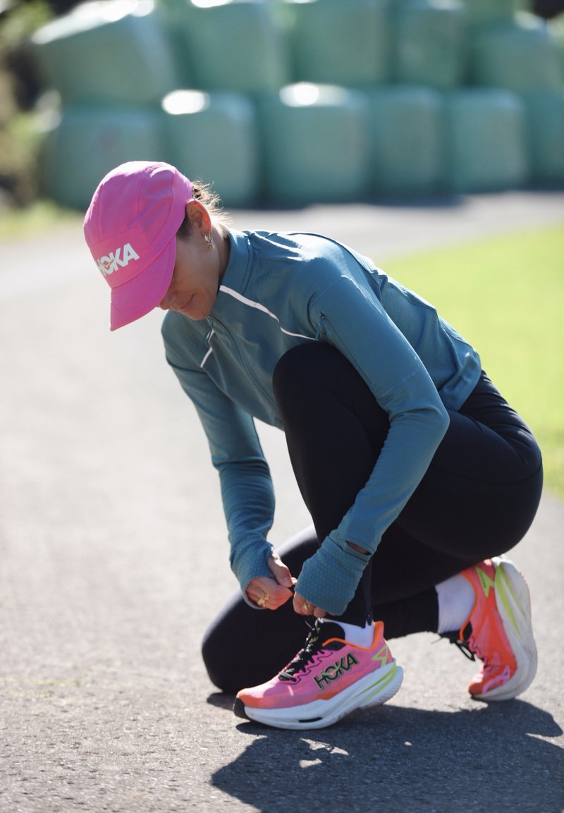Een persoon in een roze pet knielt op een betegeld pad en strikt felle roze en oranje hardloopschoenen, gekleed in een blauwe lange mouwen en zwarte leggings.