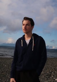 Sweat à capuche zippé bleu marine avec cordons crème, porté par-dessus une chemise blanche. En arrière-plan, une plage rocheuse et des montagnes lointaines.