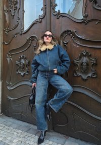 Denimist jakk kunstkarvaga kraega, kombineeritud laia säärega denimist pükstega. Mustad terava ninaosaga saapad ja väike must käekott. Puidust paneelidega taust.