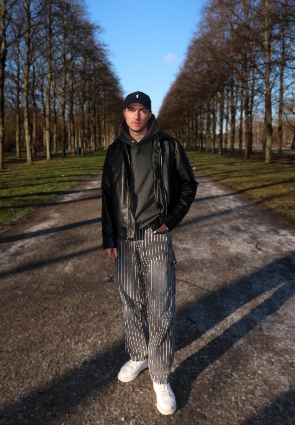 Crna kožna jakna preko sive dukserice, prugaste hlače i bijele tenisice. Pozadina prikazuje stazu sa drvećem ispod plavog neba.