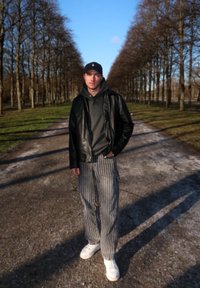 Crna kožna jakna preko sive dukserice, prugaste hlače i bijele tenisice. Pozadina prikazuje stazu sa drvećem ispod plavog neba.