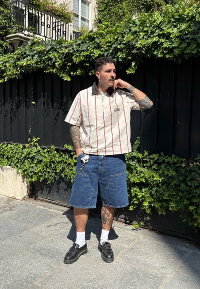 Camisa de manga corta color crema con rayas rojas y un cuello oscuro, combinada con pantalones cortos de mezclilla y zapatos negros, de pie frente a una pared cubierta de hiedra.