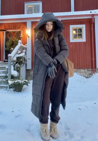 Doudoune gris foncé avec capuche oversize, associée à une combinaison marron et des bottes beige clair. Sol enneigé et marches en bois visibles.