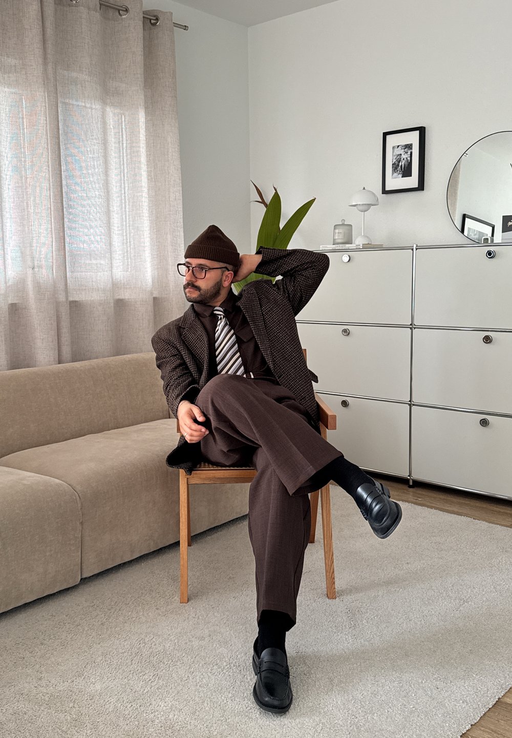 Mees pruunis ülikonnas, triibulise lipsu, prillide ja beanie'ga istub ristipidi puidust toolil modernses elutoas, kus on beež diivan ja valge kapp.