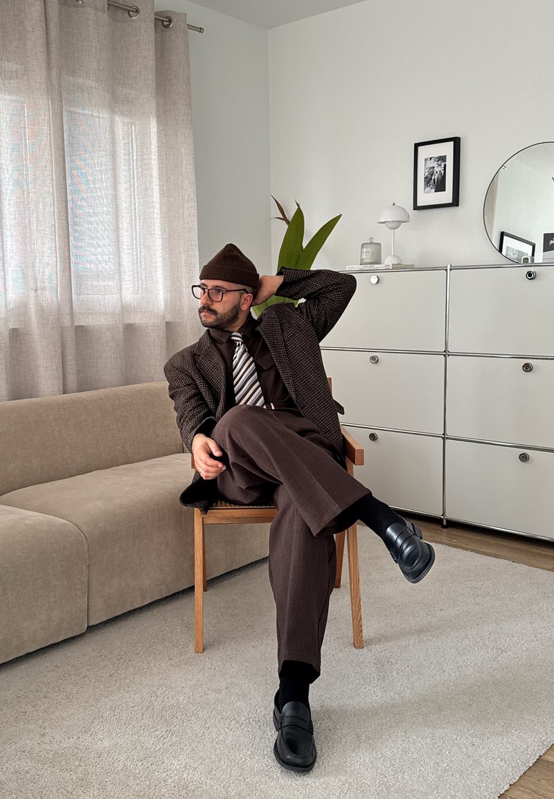 Mand iført brun jakkesæt, stribet slips, briller og beanie sidder i korslagte ben på en træstol i et moderne stue med beige sofa og hvid skab.