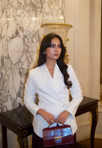 Blazer blanco ajustado con diseño envolvente, sostenido por una mujer. Ella lleva un bolso burdeos oscuro y joyas de oro. Fondo de mármol.