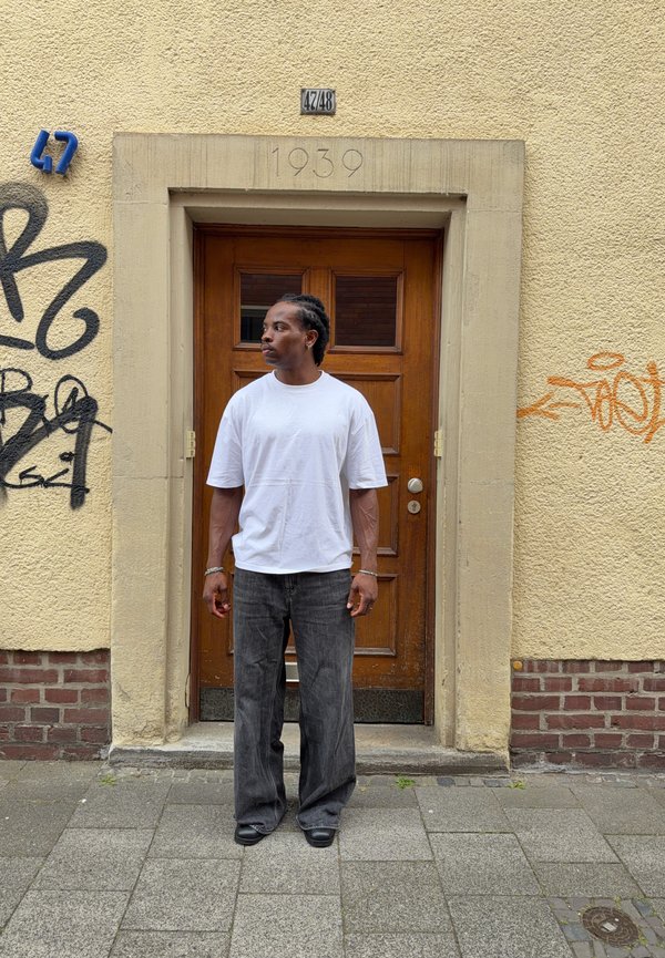 Mand i hvid T-shirt og mørke bukser står på fortovet foran en lukket trædør i en gul væg med graffiti og husnummer 47/48.