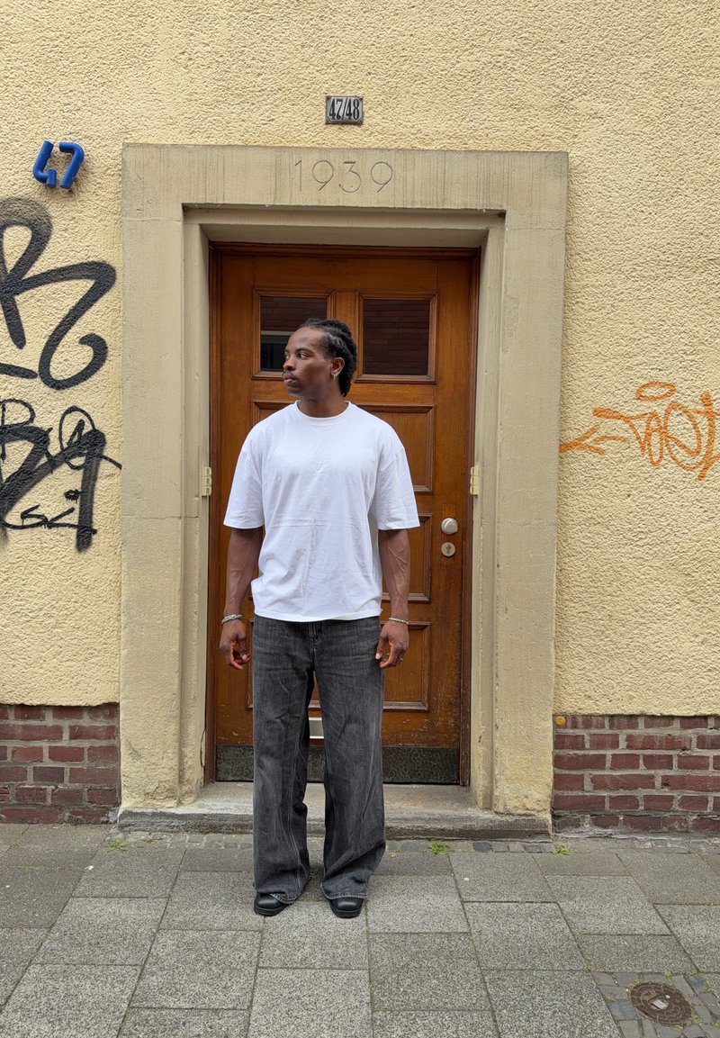 Homme en t-shirt blanc et pantalon foncé se tient sur le trottoir devant une porte en bois fermée, insérée dans un mur jaune avec des graffitis et le numéro d'adresse 47/48.