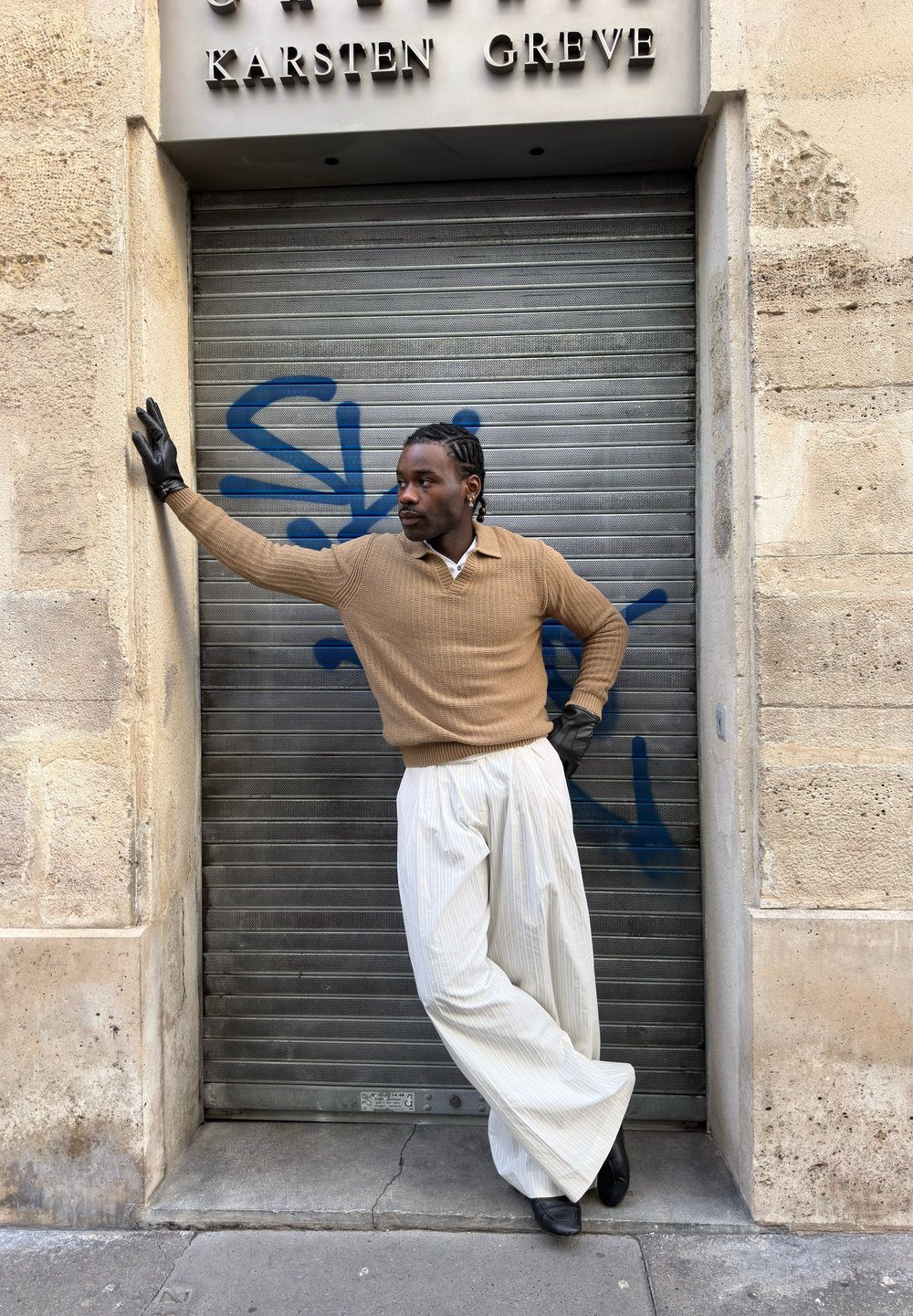 Homme aux cheveux tressés portant un pull beige, un pantalon large blanc, des gants noirs et des chaussures, appuyé contre un volet métallique couvert de graffiti.