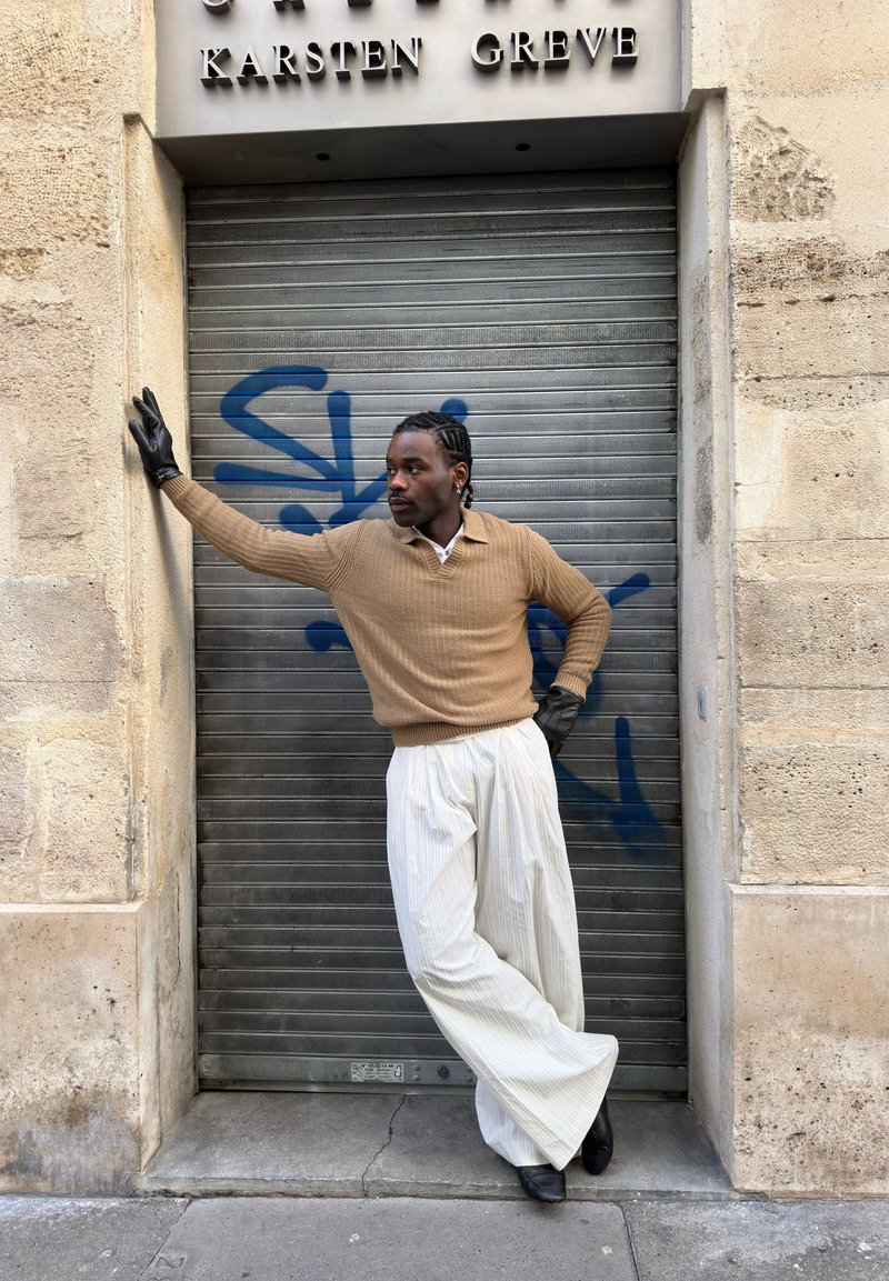 Homme aux cheveux tressés portant un pull beige, un pantalon large blanc, des gants noirs et des chaussures, appuyé contre un volet métallique couvert de graffiti.
