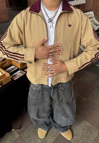 Cilvēks, kurš valkā brūnu Adidas jaku un vaļīgas pelēkas džinsus ar ķēdēm, vairākiem gredzeniem un kaklarotām, stāvot ar rokām uz krūškurvja un vēdera.