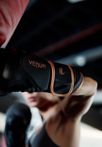 Un boxeur portant des gants Venum noirs et bruns lève le poing dans un cliché en gros plan, en action, à l'intérieur d'une salle de sport faiblement éclairée.