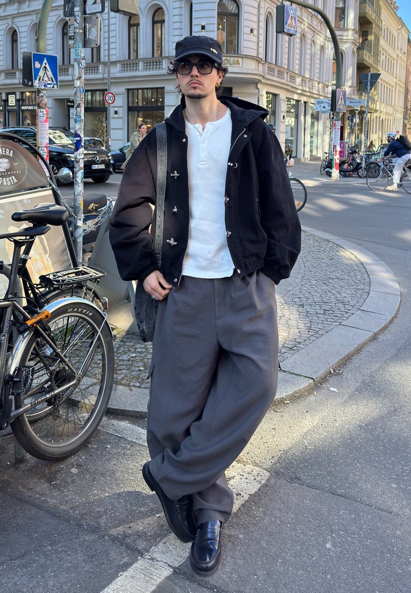 Homme portant une casquette noire, des lunettes, une chemise blanche, une veste noire et un pantalon gris ample se tient dans une rue de la ville à côté d'un vélo stationné.