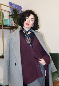 Sivý kabát, bordový kardigán s gombíkmi, čierny top a vzorovaný modrý šál. Textúrované materiály a vrstvený dizajn s vreckami.