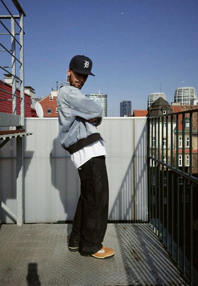 Chaqueta de denim con dobladillo acanalado negro sobre una camisa blanca, pantalones cargo negros y zapatos color crema; está de pie en un balcón con un fondo de paisaje urbano.