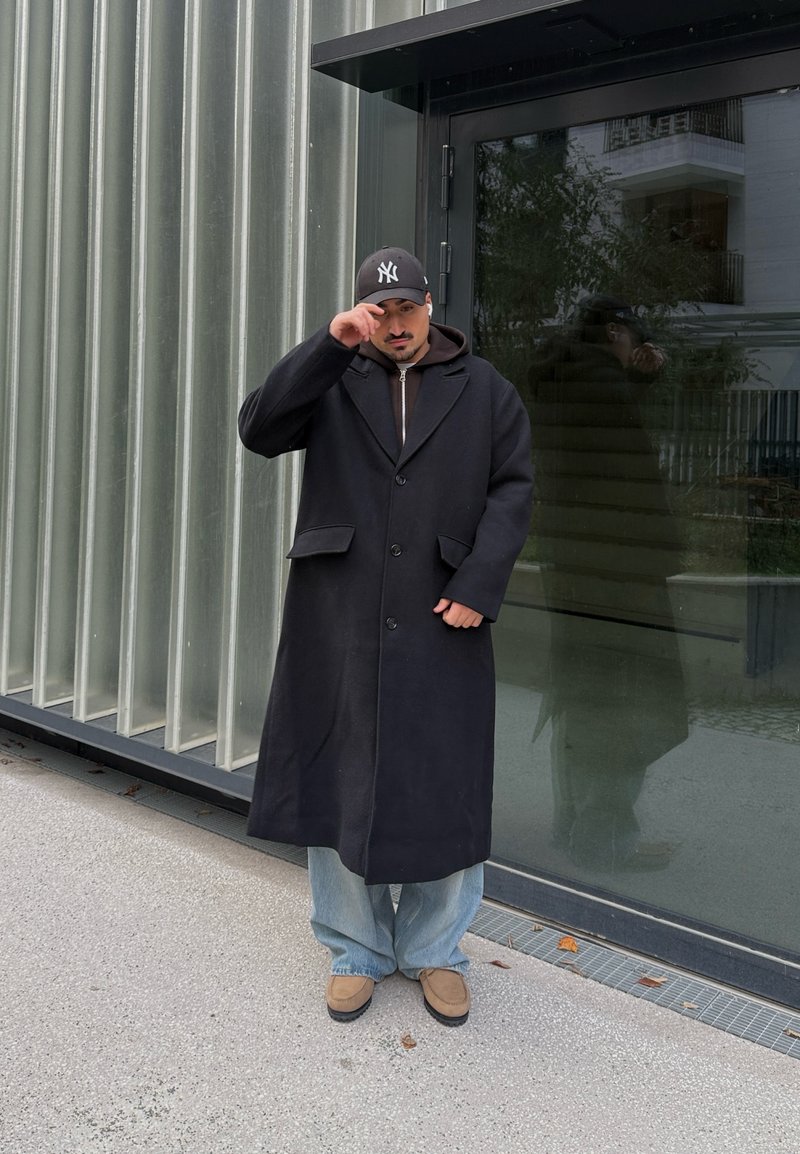 Homme portant un long manteau noir, un jean bleu et des chaussures marron, debout devant une porte en verre, ajustant une casquette noire avec un logo blanc.