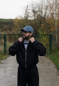 Čierna mikina na zips s voľným strihom, kombinovaná s čiernymi nohavicami. Nosená s modrou rifľovou čiapkou a sivou košeľou s modrými pruhmi pod ňou.