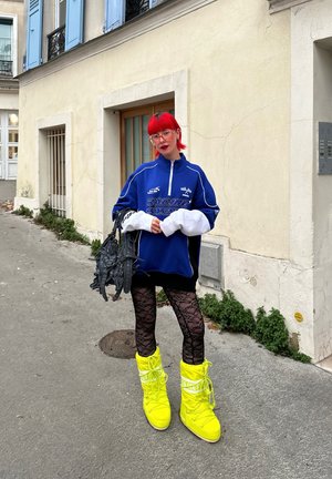 Sudadera azul de gran tamaño con cremallera y acentos blancos, leggings negros de encaje y botas amarillas hasta la rodilla. Sostiene un bolso negro texturizado.
