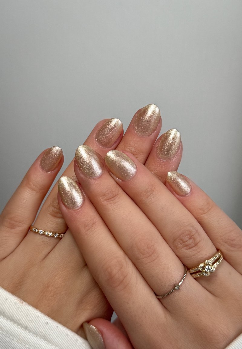 Vernis à ongles en or métallique avec une finition brillante, présentant des formes amandes. Deux bagues argentées et une bague dorée aux doigts. Texture lisse.