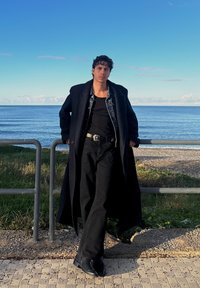 Long manteau noir sur une chemise noire et une veste en denim, associé à un pantalon et des chaussures noirs. Debout près d'une rambarde avec un paysage marin en arrière-plan.