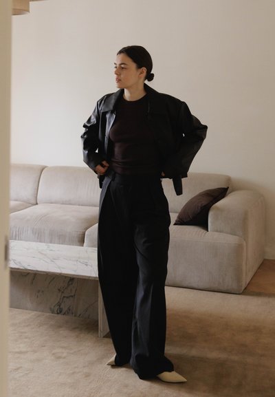 Chaqueta de cuero negra sobre una camiseta marrón oscuro, combinada con pantalones negros holgados y zapatos puntiagudos beige. Sofá de tonos neutros en el fondo.