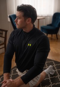 T-shirt de sport noir à manches longues avec un col zippé et un logo vert, porté avec un short blanc. La personne est dans une position d'étirement assise.