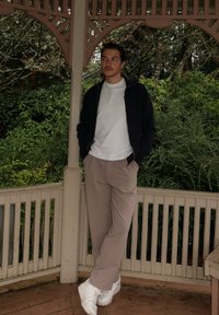 Tenue décontractée comprenant une veste à zip noire, un t-shirt blanc, un pantalon beige et des baskets blanches, sur un fond de feuillage vert.
