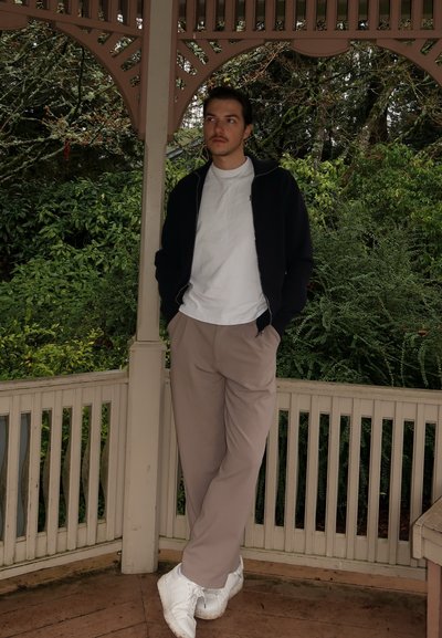 Conjunto casual que incluye una chaqueta negra con cremallera, camiseta blanca, pantalones beige y zapatillas blancas, con un fondo de follaje verde.