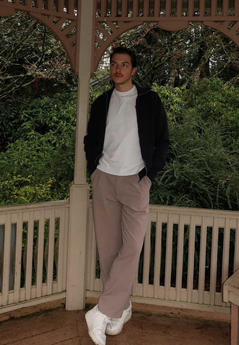 Casual outfit met een zwart jack met ritssluiting, een wit T-shirt, beige broek en witte sneakers, tegen een achtergrond van groene bladeren.