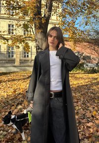 Abrigo largo negro sobre una camiseta blanca, combinado con vaqueros oscuros y un cinturón negro. A su lado, un perro pequeño con correa está de pie en un parque cubierto de hojas.