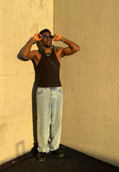 Top sin mangas negro, jeans holgados azul claro con texto blanco en el costado, gafas de sol negras y zapatos negros con suela gruesa frente a una pared gris.