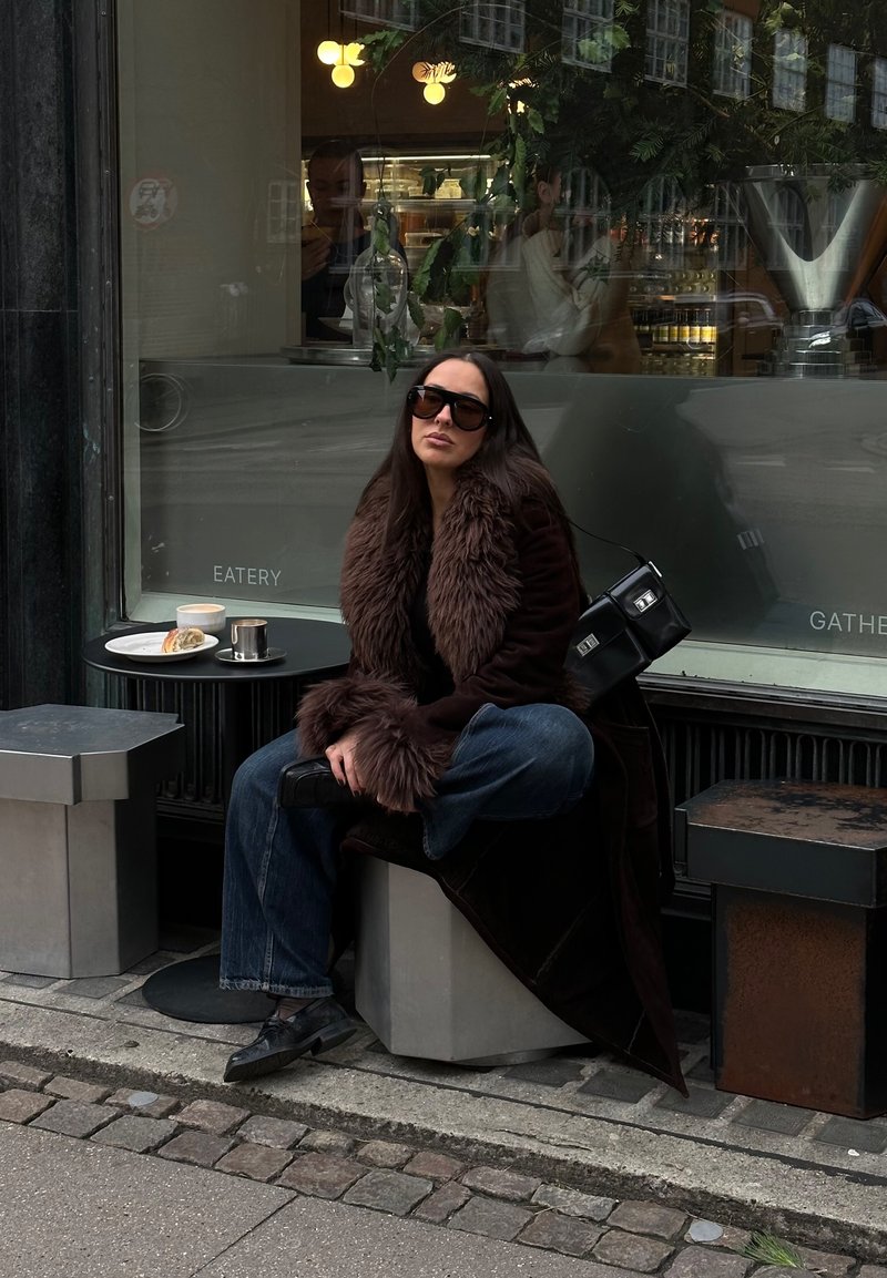 Frau in dunklem Pelzmantel und Sonnenbrille sitzt an einem Café-Tisch im Freien mit Kaffee und Gebäck an einem bewölkten Tag.